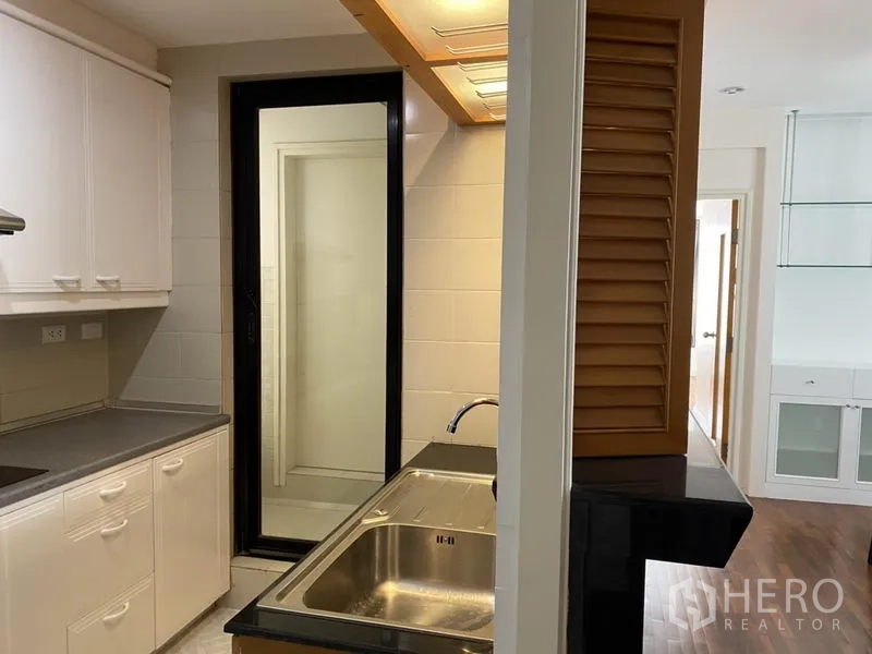 Condominium for rent or sale in Sathon, Bangkok - Galley kitchen with stainless sink, white cabinets and a sliding glass door to a utility balcony.