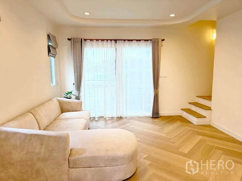 Townhouse for rent in Suan Luang, Bangkok - Bright living room with an L-shaped beige sofa, herringbone wood floors, sheer-draped sliding doors, and a short staircase.