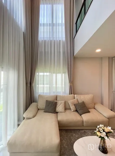 Detached House for rent or sale in Bang Phli, Samut Prakan - L-shaped sofa in a double-volume living room with full-height windows and mezzanine railing.