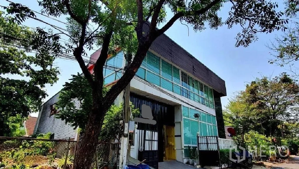 Home Office for rent or sale in Bang Khun Thian, Bangkok - Contemporary facade with green-tinted glass, gated entry, and mature trees.