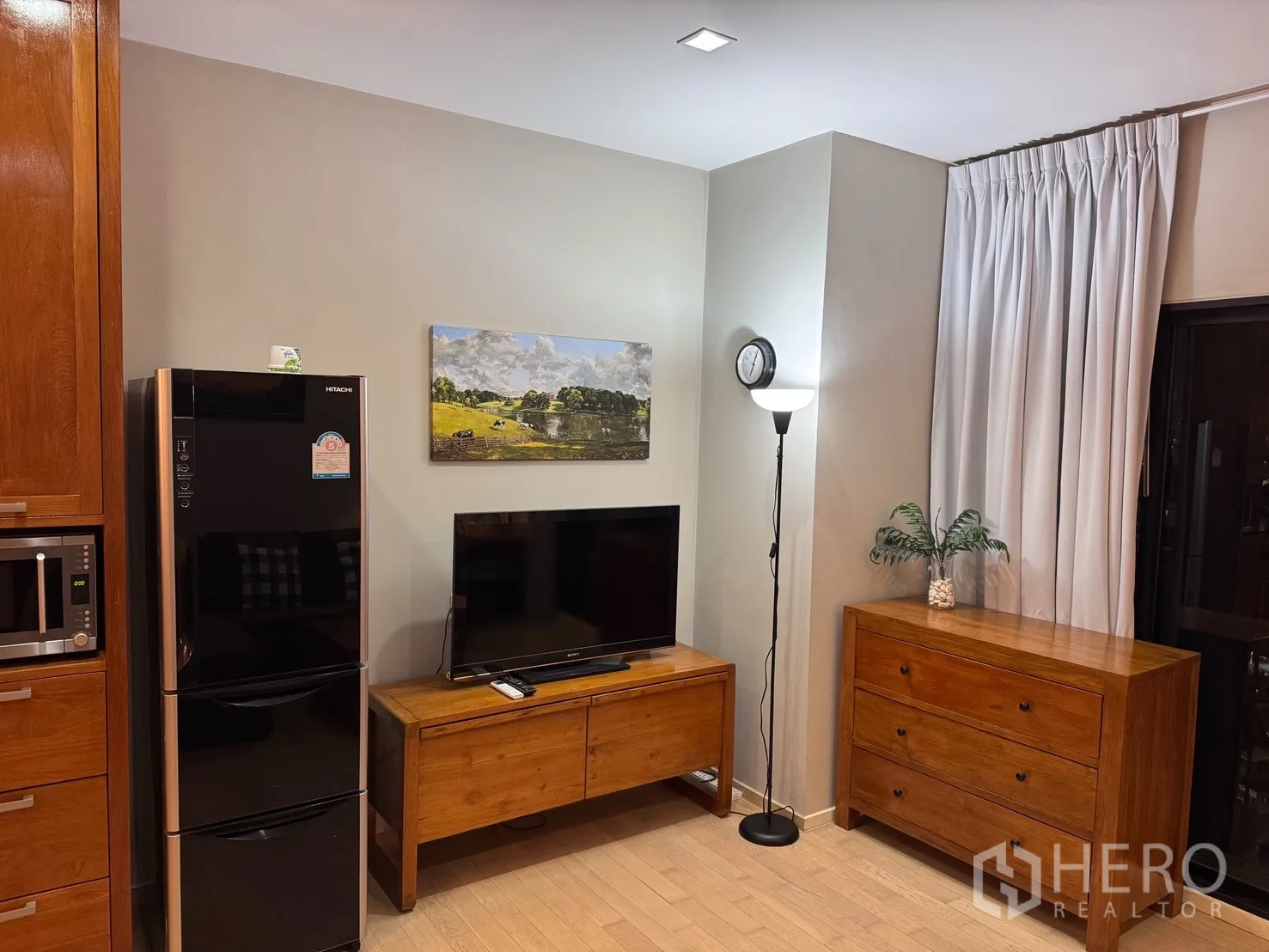 公寓 for rent or sale in Watthana, Bangkok - 电视机摆放在木质电视柜上，旁有黑色冰箱、抽屉柜与落地灯，窗帘素雅。