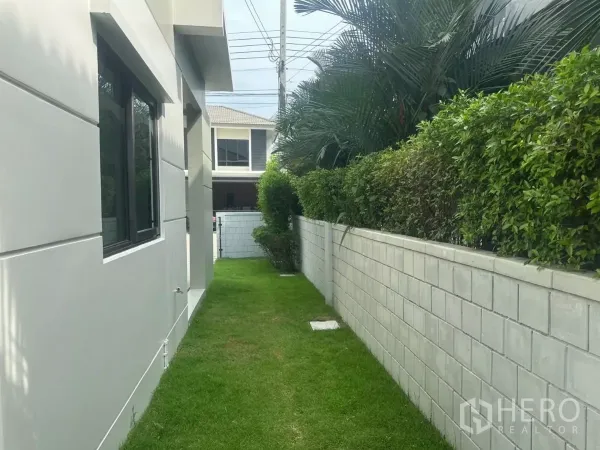 独立式住宅 出售在Samut Prakan - 侧院草坪步道，一侧为绿篱与围墙，另一侧为房屋窗户。