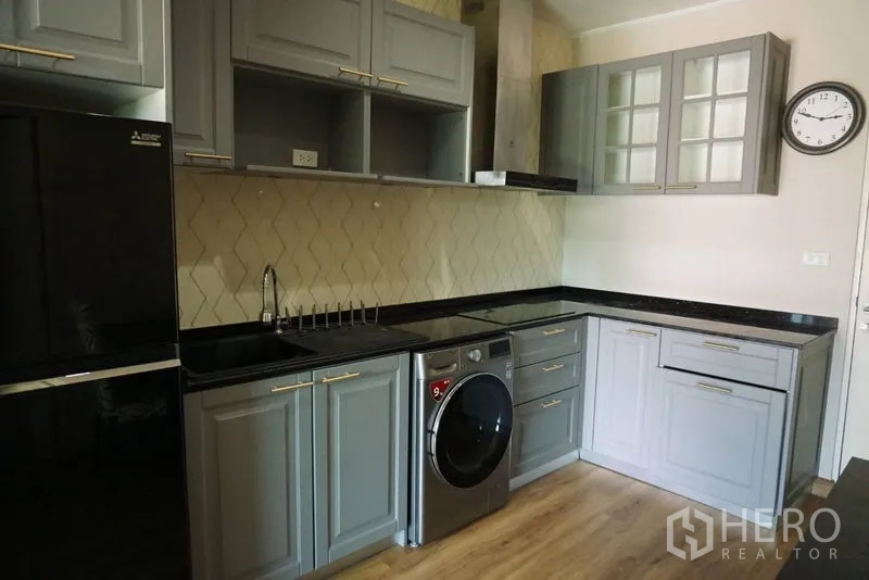 Condominium for sale in Khlong Toei, Bangkok - Close-up of L-shaped kitchen showing shaker-style cabinets, cooktop, hood and washing machine.