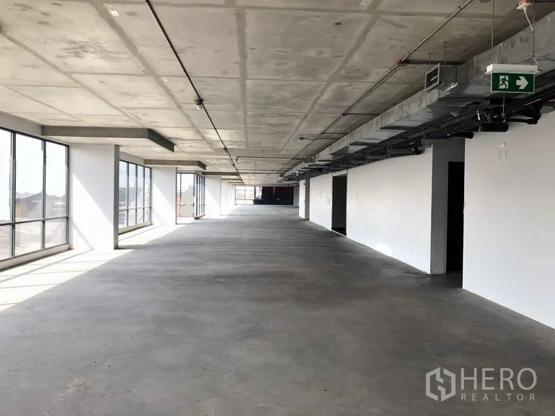 Office Space for rent in Suan Luang, Bangkok - Long open office floor with concrete slab ceiling, emergency exit sign, and continuous windows along one side.