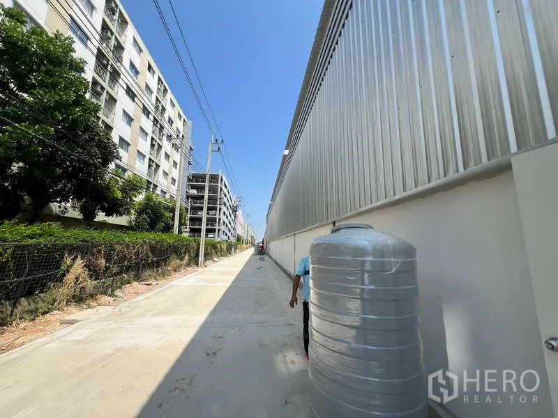 货栈 出租在Lat Krabang, Bangkok - 金属外墙仓库旁的混凝土通道，沿线有公寓与电线杆，晴空下的叻甲挽街景。