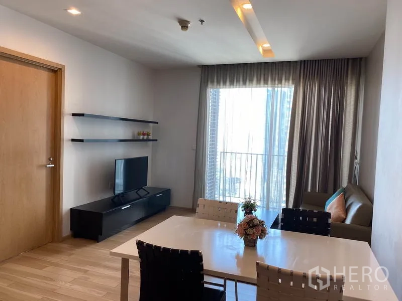 Condominium for rent in Khlong Toei, Bangkok - Dining area with four-seater table near the balcony and TV cabinet, sheer curtains filtering light.
