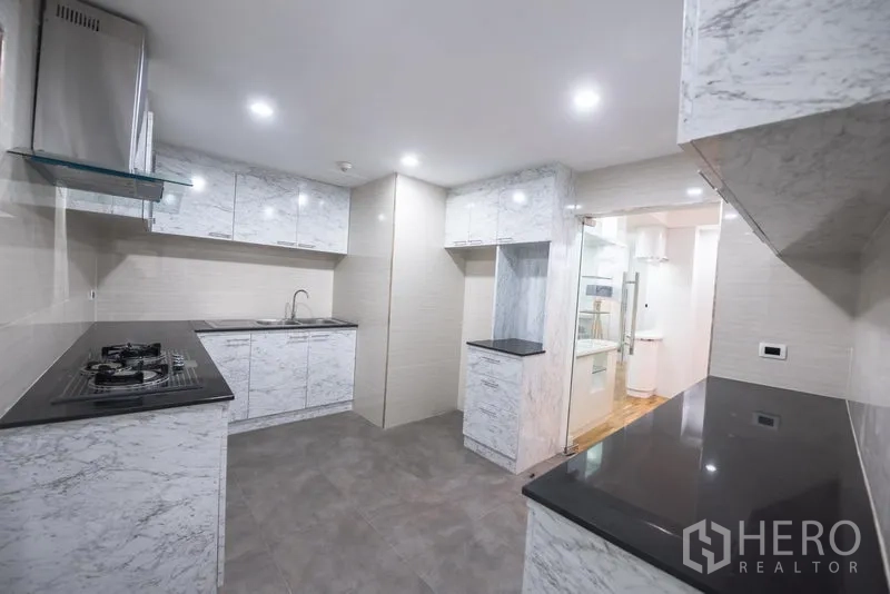 Condominium for rent in Khlong Toei, Bangkok - Modern L-shaped kitchen with marble cabinets, black worktops, recessed lighting and doorway to living area.