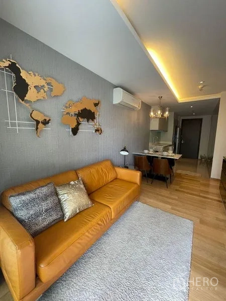Condominium for rent in Watthana, Bangkok - Living room with tan leather sofa, accent wall map art, rug, and sliding doors to the balcony.