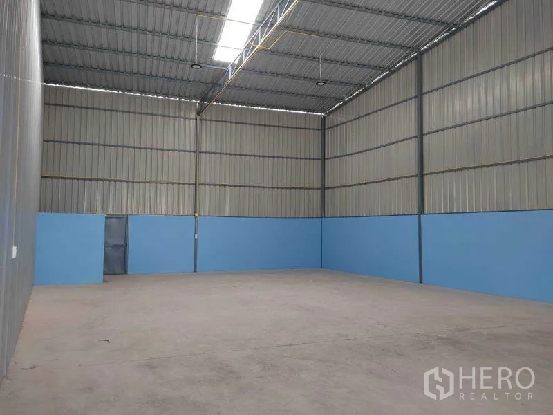 Warehouse for rent in Bang Yai, Nonthaburi - Empty warehouse interior with high metal ceiling, skylight strip, blue lower walls, and smooth concrete floor.