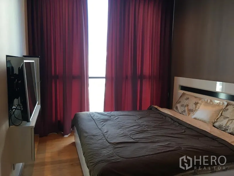Condominium for rent or sale in Watthana, Bangkok - Bedroom with large window, wall-mounted TV opposite the bed and red curtains.