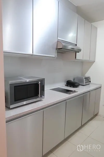 Condominium for rent in Khlong Toei, Bangkok - Galley kitchen with metallic cabinetry, electric cooktop, range hood, microwave and toaster.