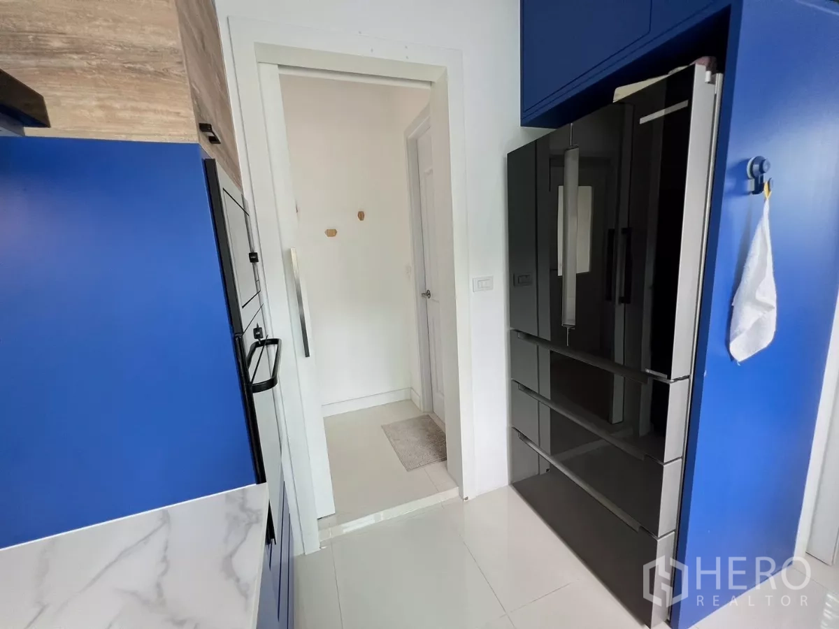 Detached House for rent or sale in Khlong Luang, Pathum Thani - Kitchen entry showing large black refrigerator within blue cabinetry.