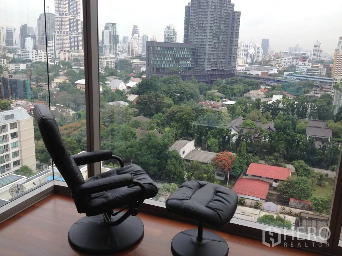Condominium for rent in Khlong Toei, Bangkok - Corner glass window with lounge chair and ottoman facing green city views.