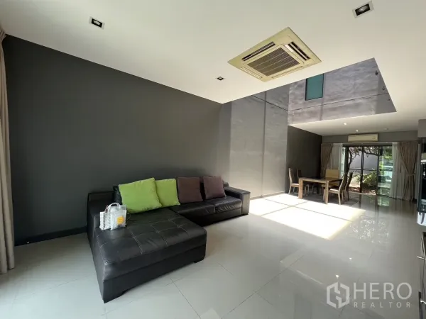 Townhouse for rent in Lat Phrao, Bangkok - Bright living and dining area with double-height ceiling, large windows and black leather sofa.